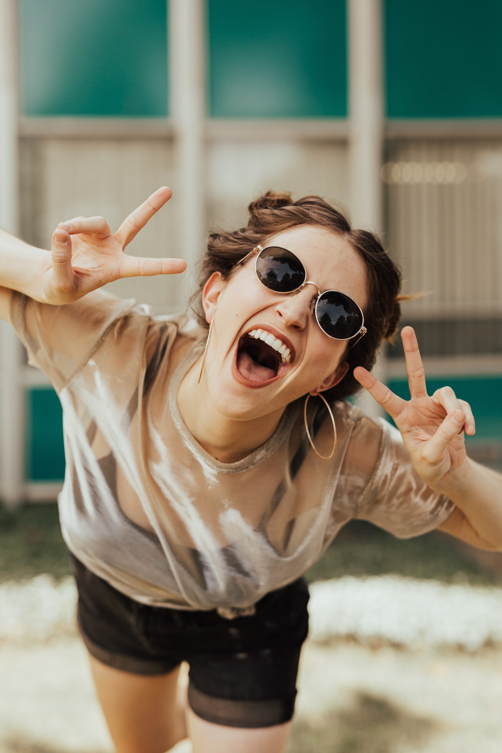 Young woman in trendy sunglasses laughing at camera with double peace signs