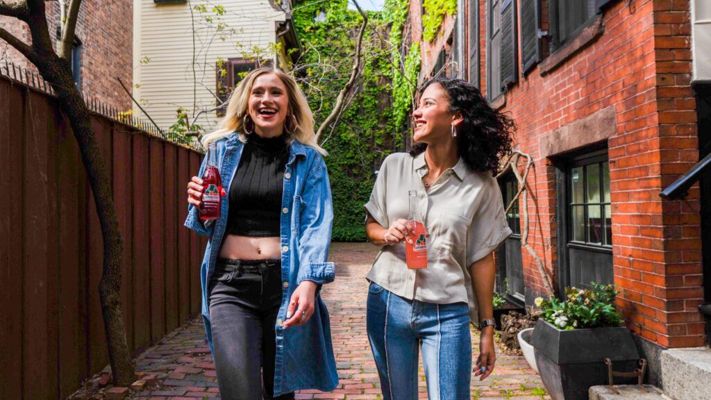 Two 20-something women laughing whilst walking in the sun and drinking soda