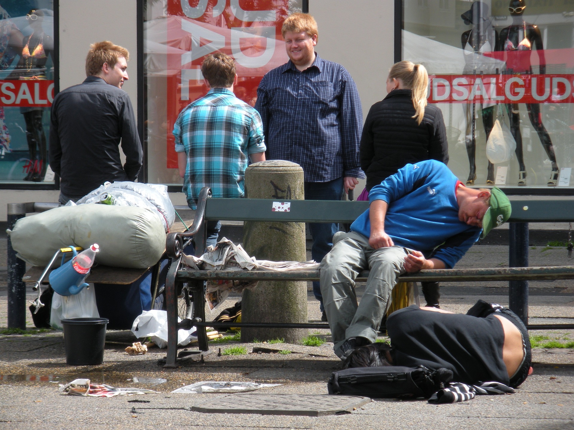 A drunk man sleeps on a bench whilst people laugh in the background