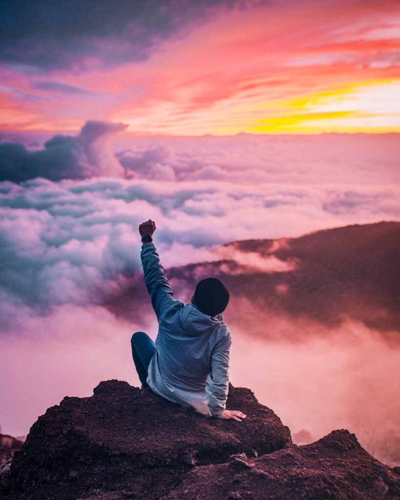 A young man punching the sky in exaltation. He is sitting on a mountain surrounded by pink clouds