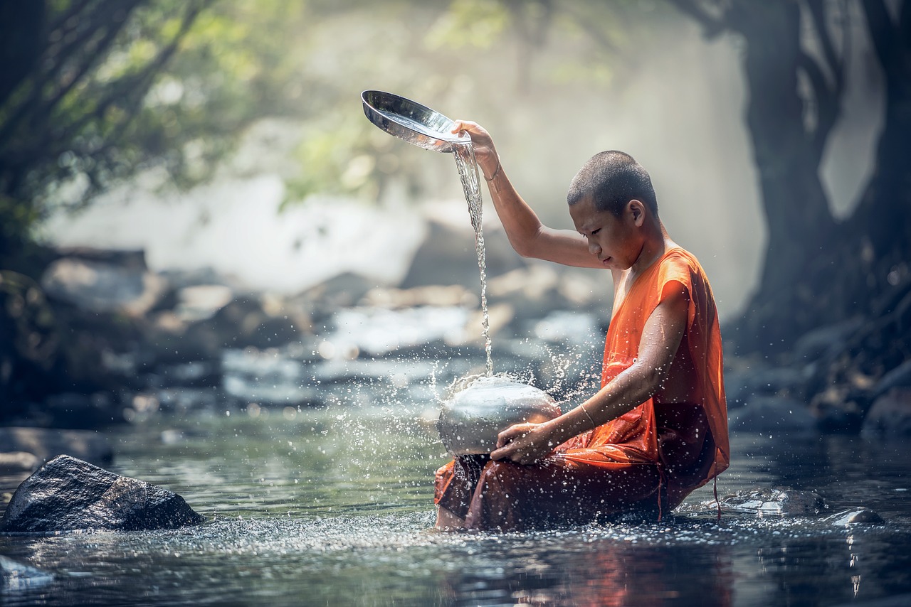 A young monk in saffron robes pouring water from a pan into creek